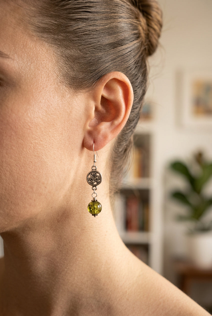 A pair of handmade dangle earrings featuring oxidized silver circular charms with a Celtic knot design, connected to silver fishhooks and finished with olive green glass beads, shown on a light gray background.