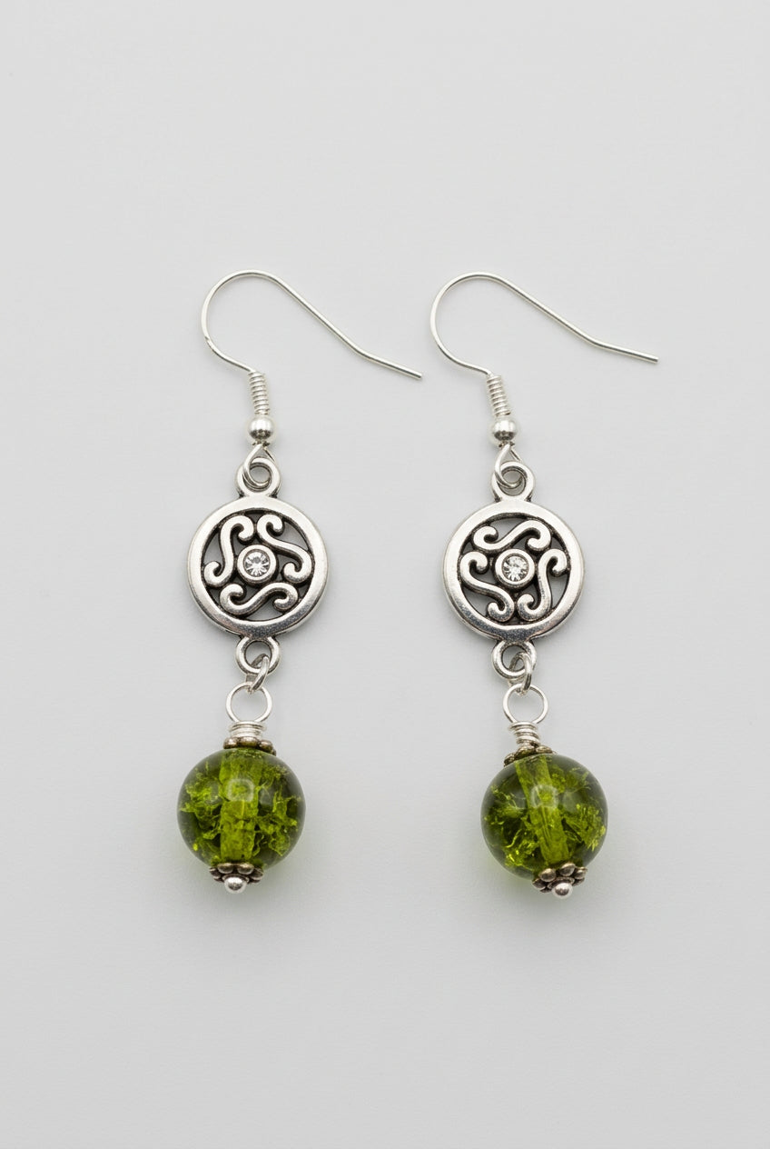 A pair of handmade dangle earrings featuring oxidized silver circular charms with a Celtic knot design, connected to silver fishhooks and finished with olive green glass beads, shown on a light gray background.