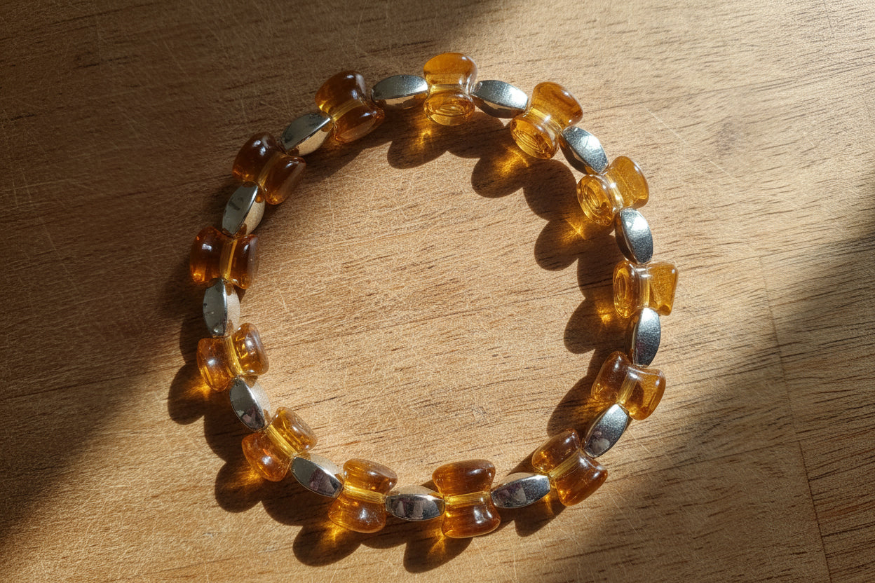 A beaded stretch bracelet made of translucent amber-colored beads separated by polished silver-toned metal beads, displayed on a wooden surface with dappled sunlight and shadows.