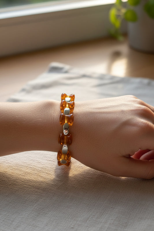 A close-up of a person's wrist wearing a translucent amber-colored beaded bracelet with small silver-toned spacer beads, resting on a light fabric surface near a window.