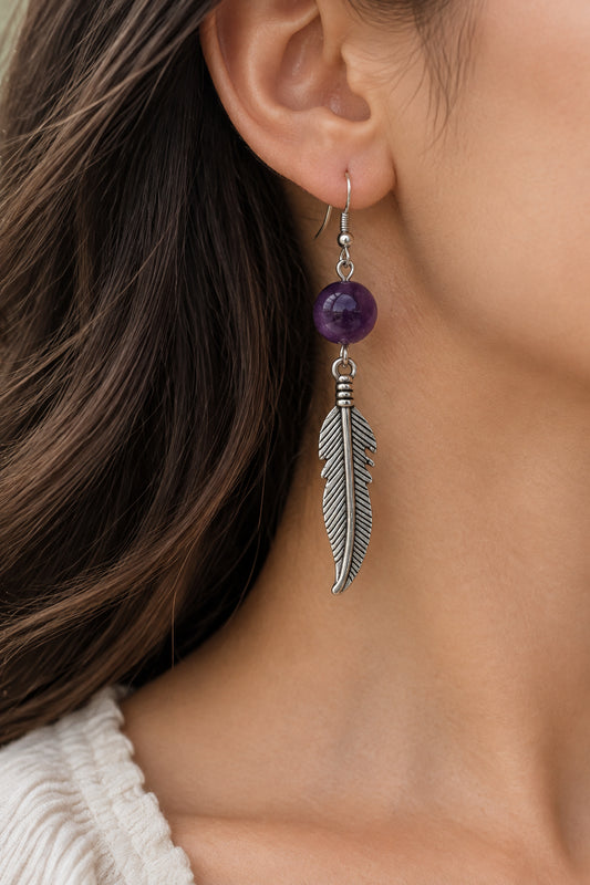 A pair of silver-toned dangle earrings with round purple beads and feather-shaped charms, displayed on a light gray surface.