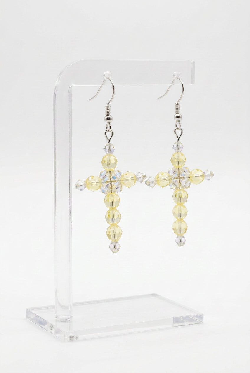 A pair of handmade cross dangle earrings made of faceted beads, featuring pale yellow beads and clear beads, hanging from silver fishhook wires on a light gray background.