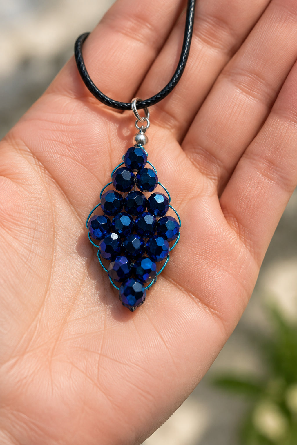 A beaded pendant necklace featuring dark blue faceted beads arranged in a teardrop shape, suspended from a black cord with silver-toned metal findings, set against a textured off-white background.
