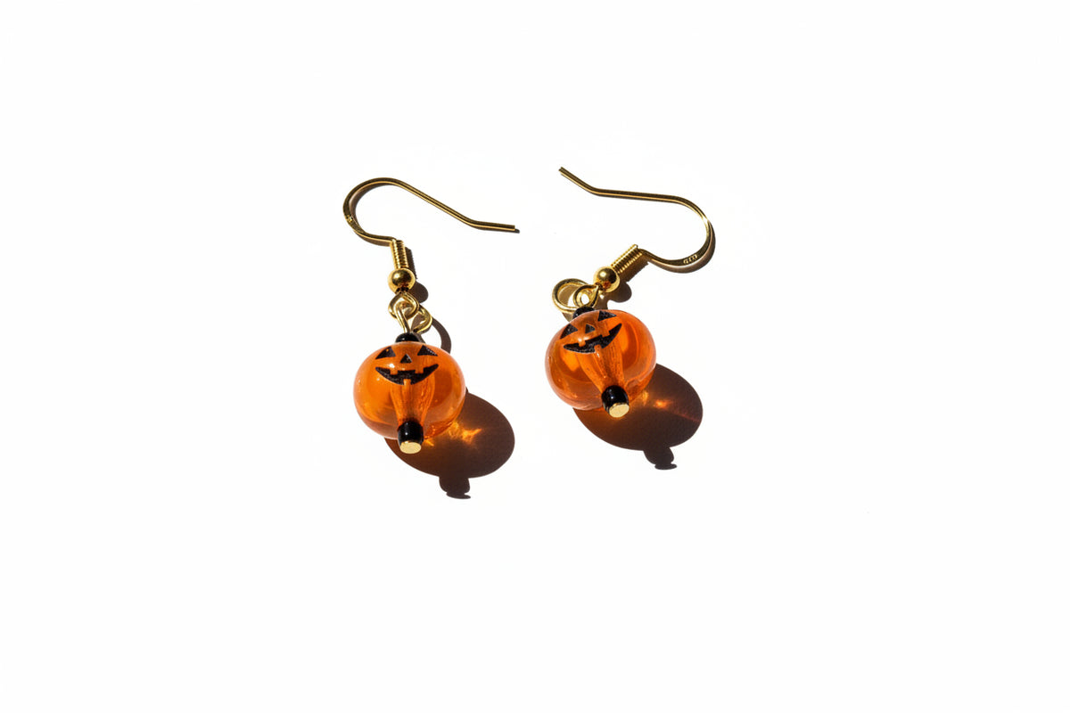 A pair of gold-tone dangle earrings with translucent orange pumpkin-shaped beads featuring black jack-o'-lantern faces, displayed on a textured white fabric background.