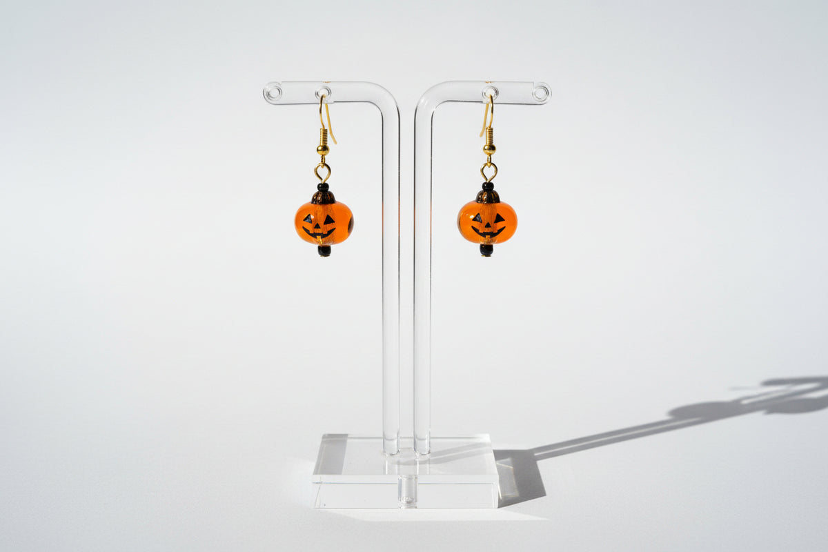 A pair of gold-tone dangle earrings featuring orange translucent pumpkin-shaped beads with black jack-o'-lantern faces and dark brown teardrop-shaped beads behind them, displayed on a textured white fabric background.