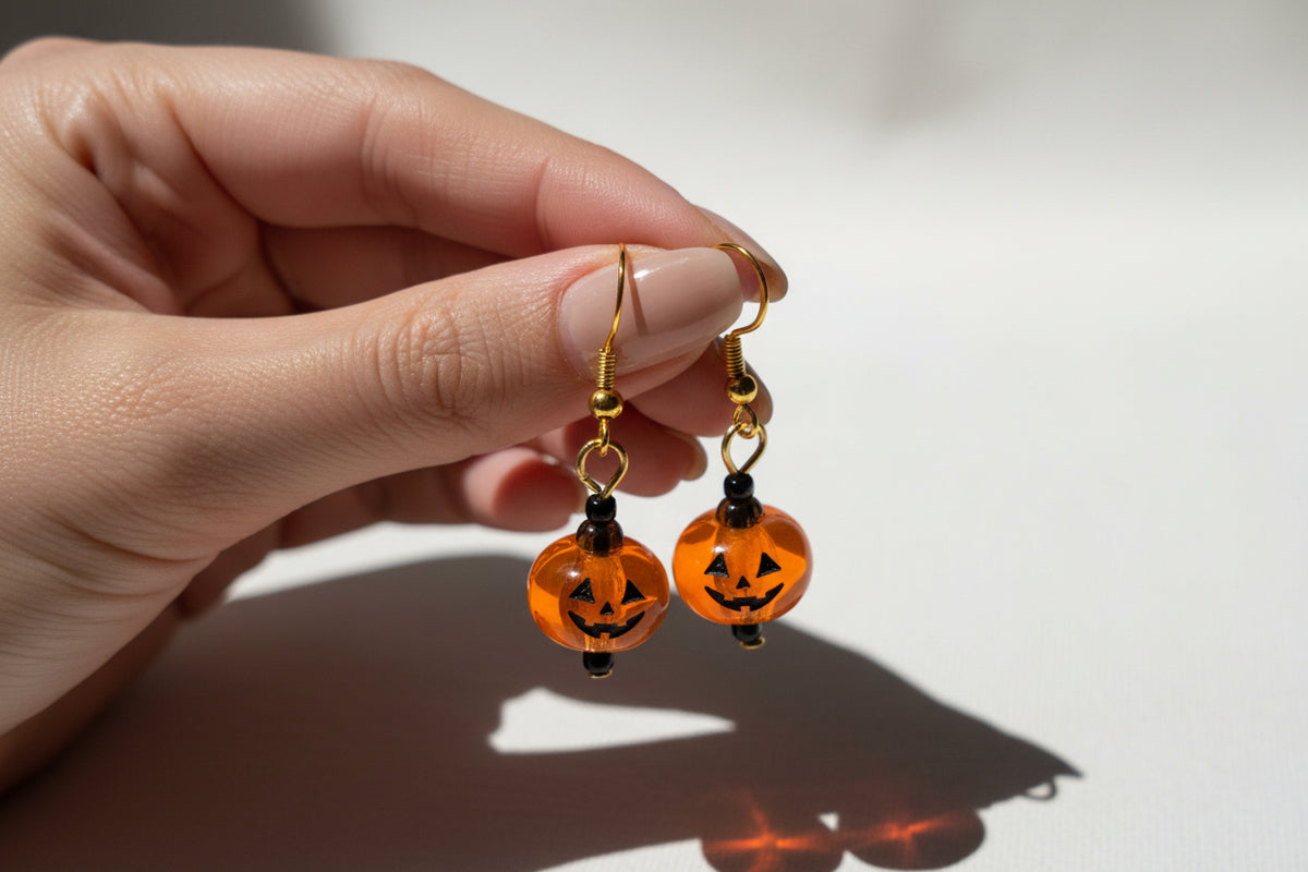 A pair of gold-tone dangle earrings with translucent orange pumpkin-shaped beads featuring black jack-o'-lantern faces, displayed on a textured white fabric background.