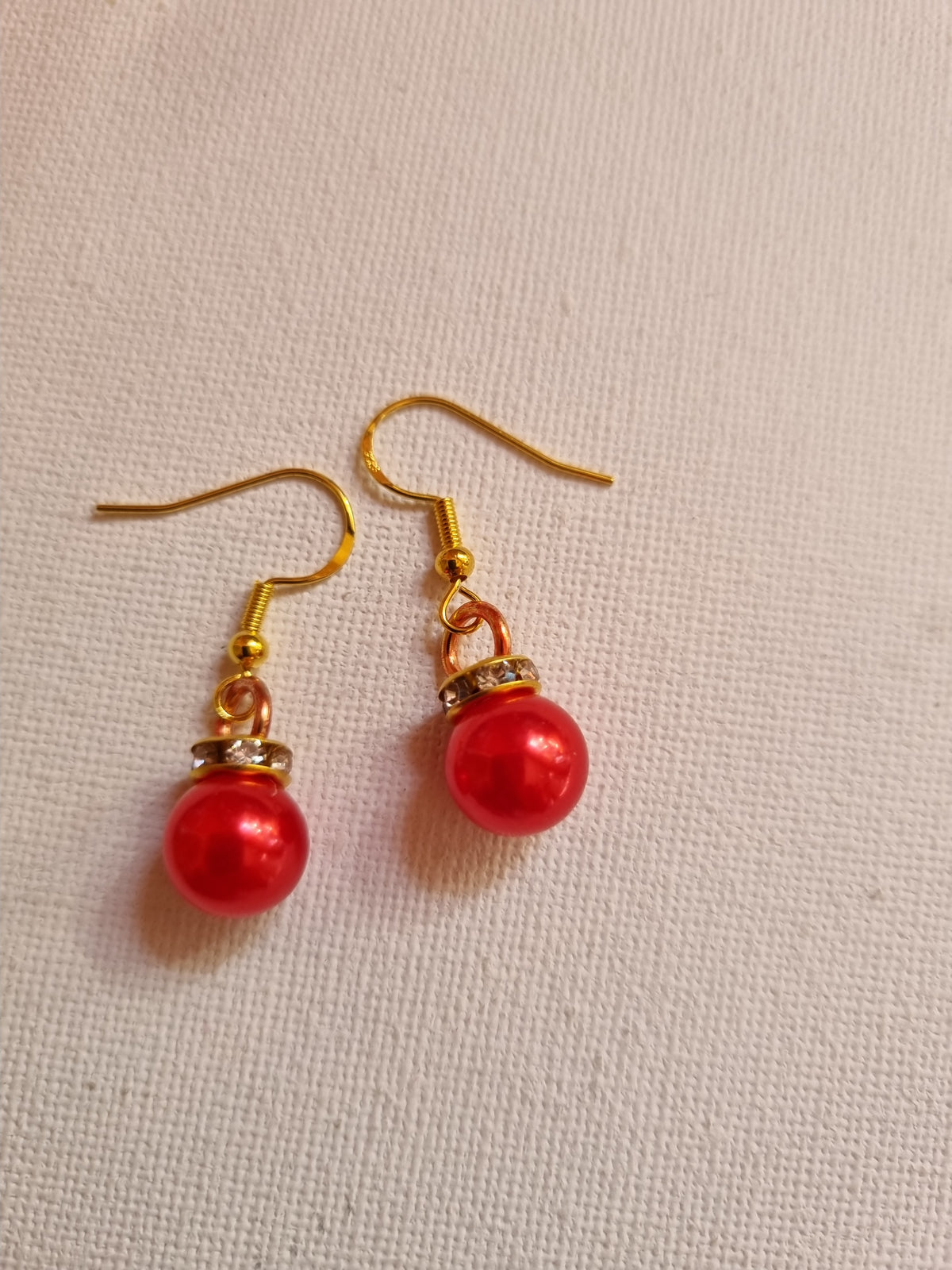 A pair of gold-tone dangle earrings with glossy red pearl-like beads and small gold spacer details, displayed on a textured off-white fabric background.
