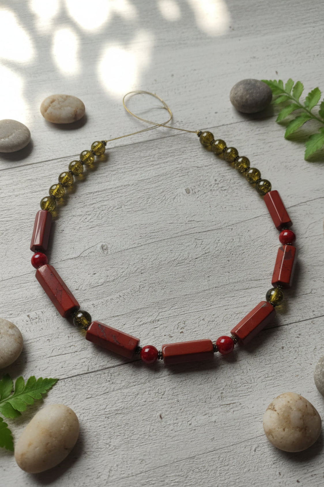 Necklace with red and green beads on a light wooden surface with stones and leaves.