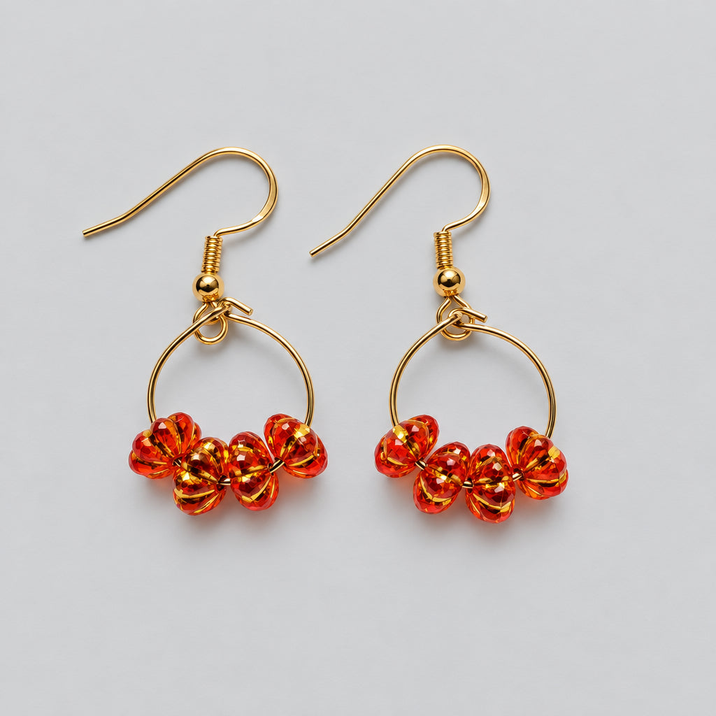 A pair of gold-tone dangle earrings with small circular hoops and clusters of red-orange faceted beads at the bottom, shown on a textured white fabric background.