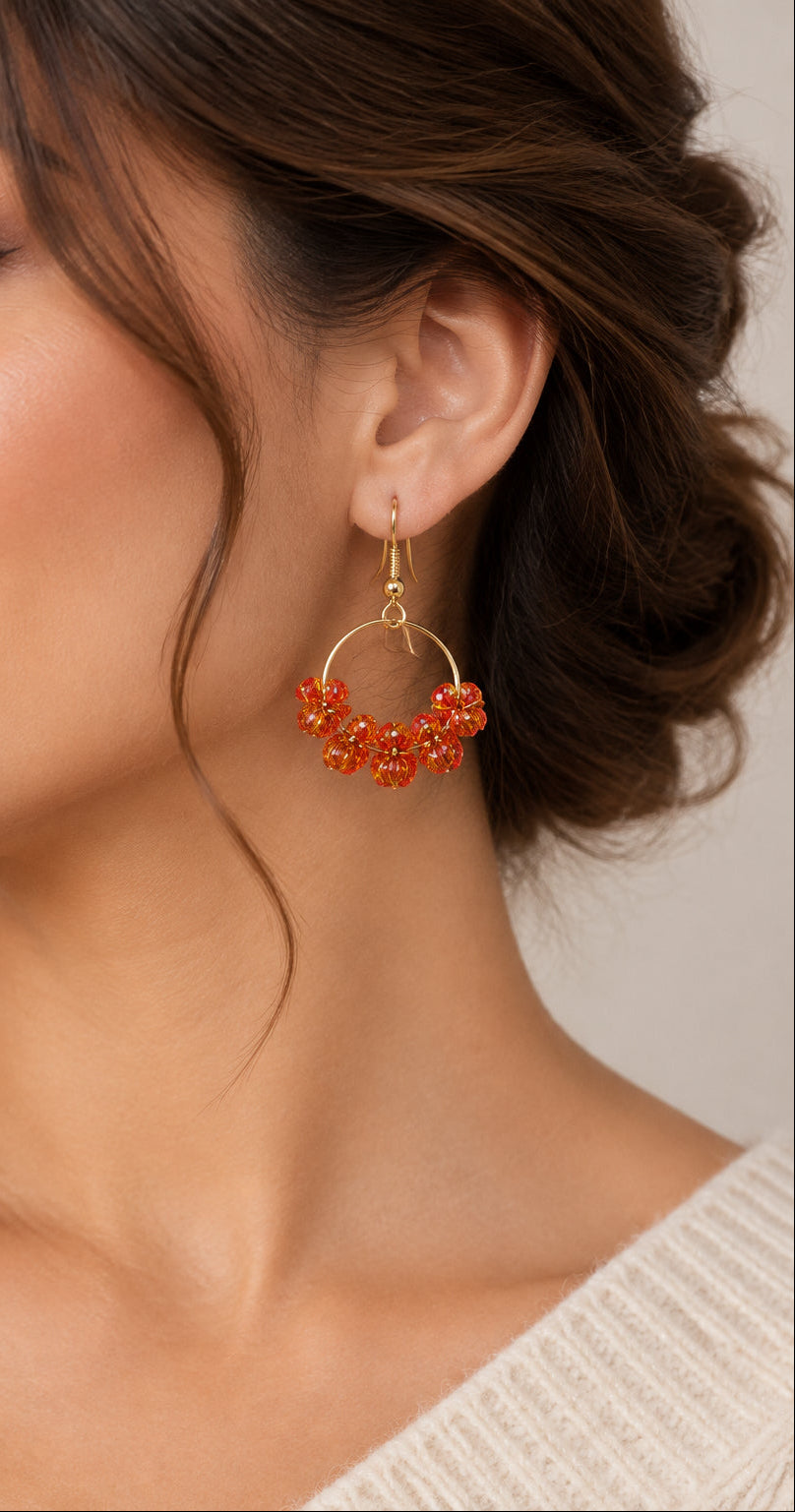 A pair of handmade dangle earrings featuring gold-tone metal hoops and fishhook wires, adorned with clusters of small red-orange translucent beads at the bottom, displayed on a textured white fabric background.