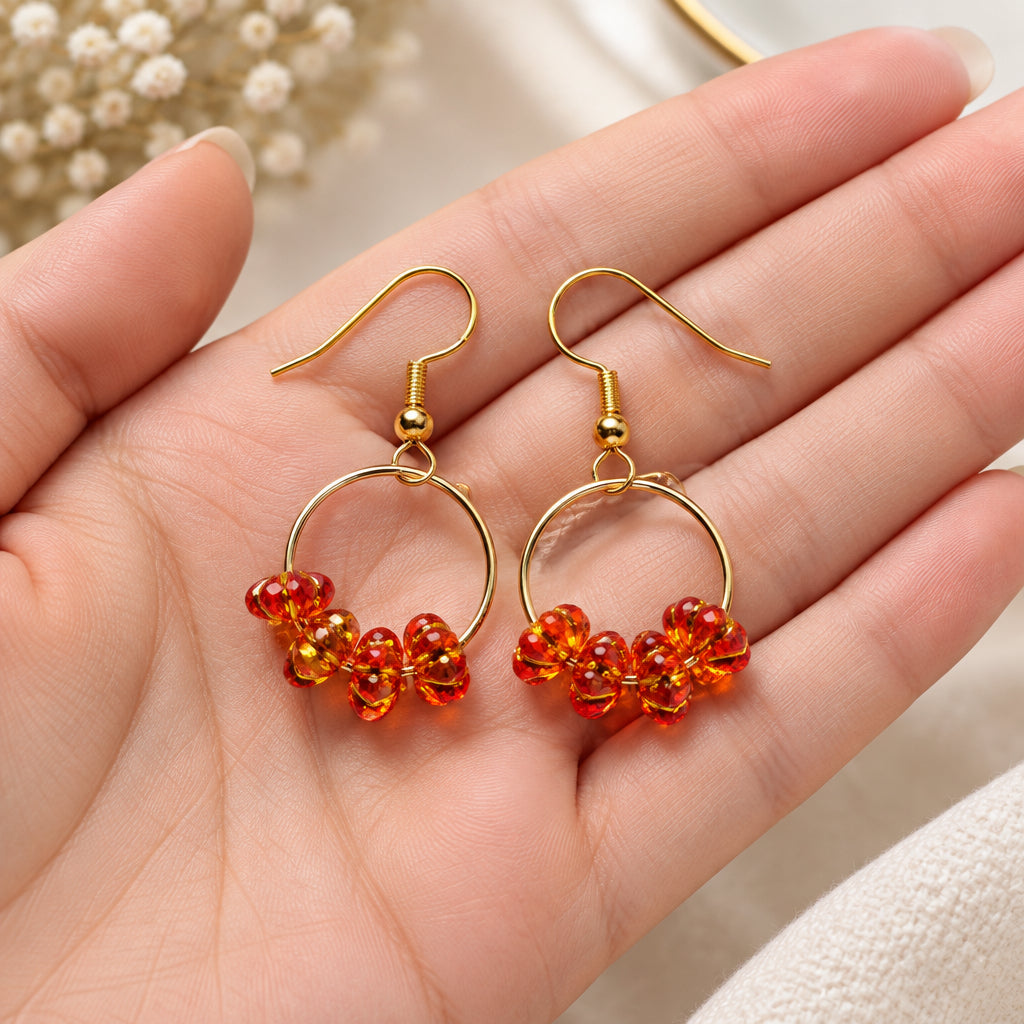 A pair of handmade dangle earrings featuring gold-tone metal hoops and fishhook wires, adorned with clusters of small red-orange translucent beads at the bottom, displayed on a textured white fabric background.